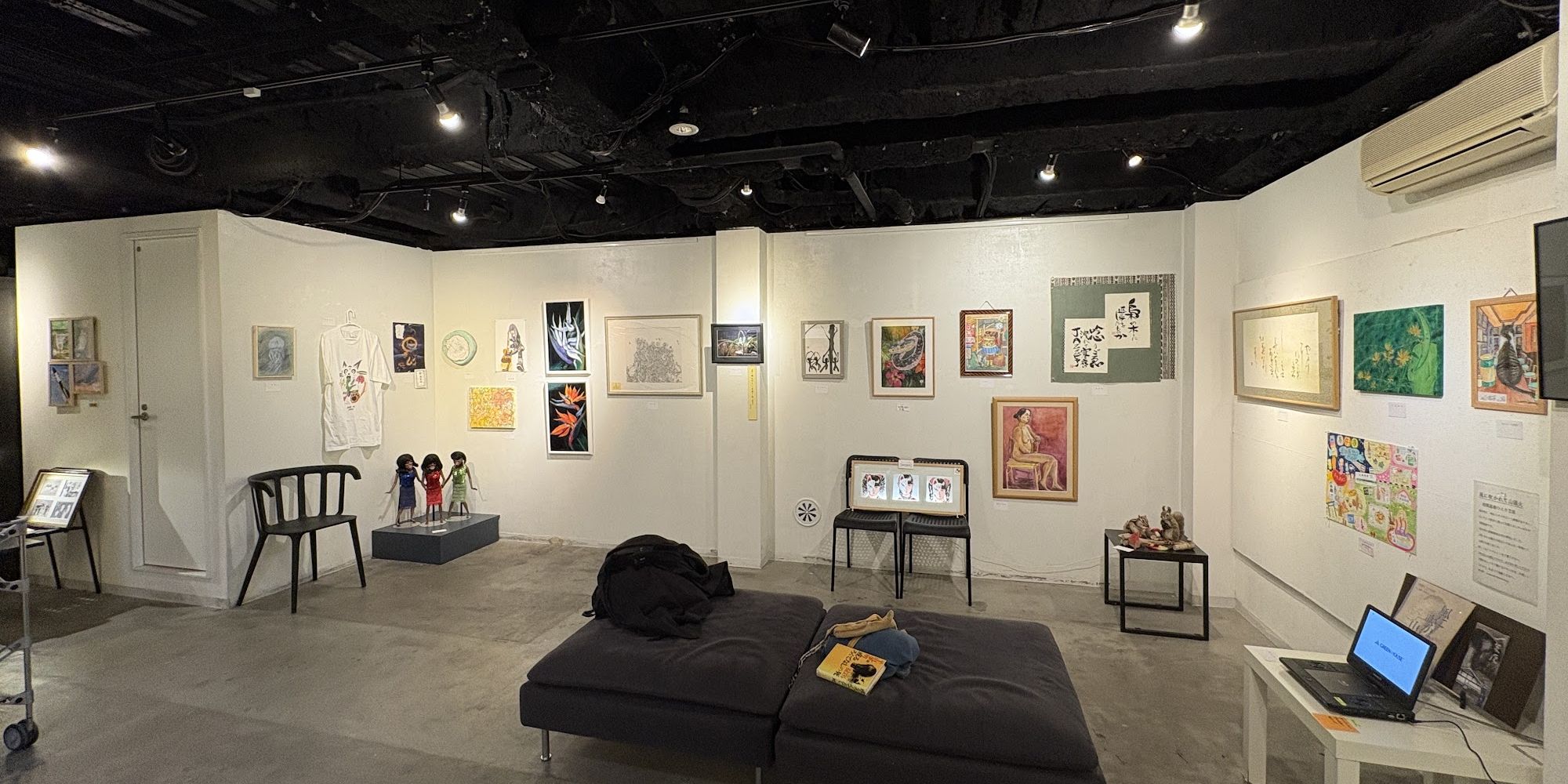展示中のギャラリーの全景。白い壁に絵画が並び、黒い天井からスポットライトが当たっている